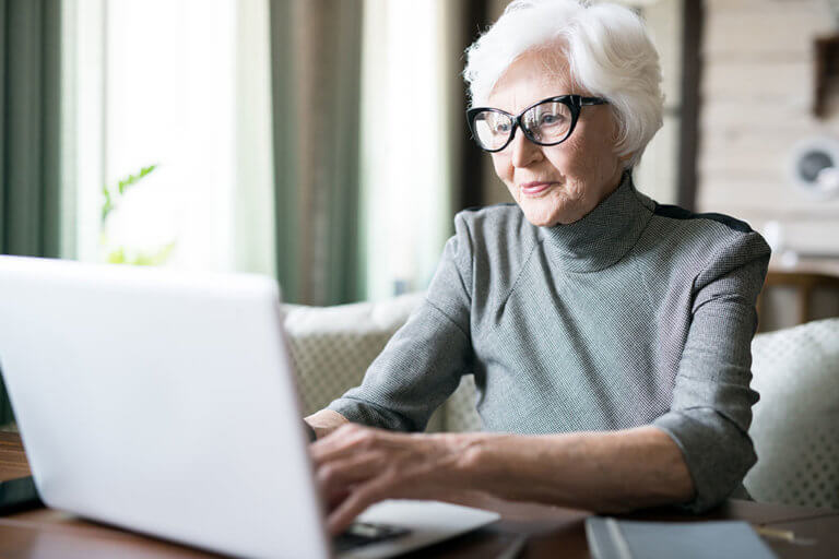 White haired senior woman wearing eyeglasses sitting at cafe and using her laptop