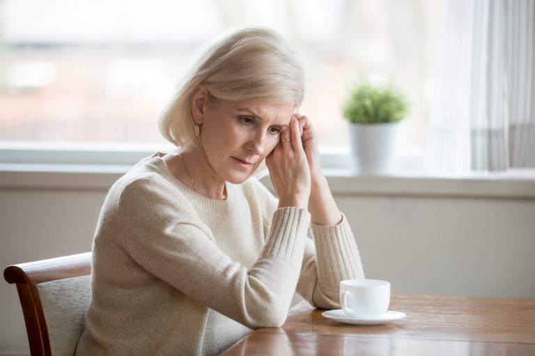 Concerned aged woman sit at table lost in thoughts