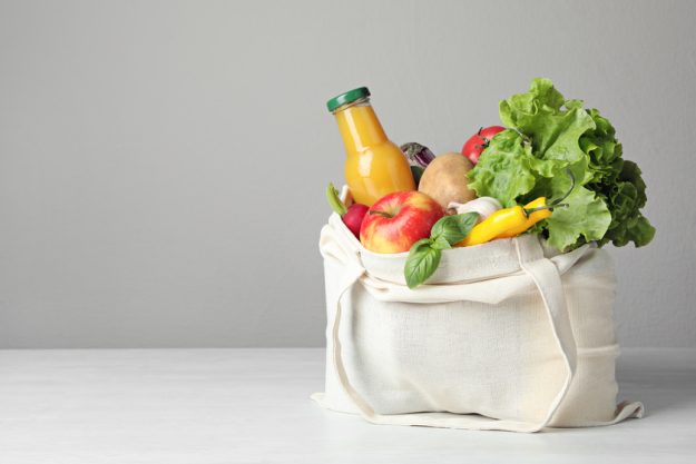 Cloth bag with vegetables and bottle of juice on table against g