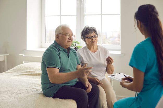 Worried senior couple sitting on bed and talking to nurse or family practitioner Worried senior couple sitting on bed and talking to nurse or family practitioner