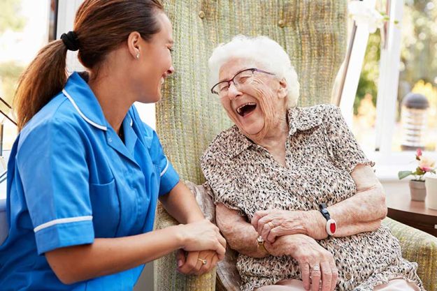 Senior Woman Sitting In Chair And Laughing With Nurse In Retirement Home