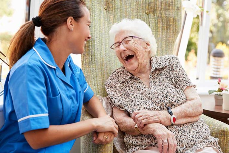 Senior Woman Sitting In Chair And Laughing With Nurse In Retirement Home