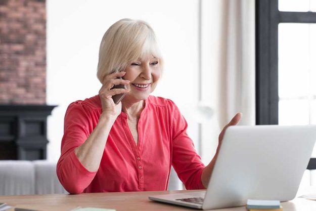 Smiling senior old elderly businesswoman talking on mobile phone while working at home with laptop