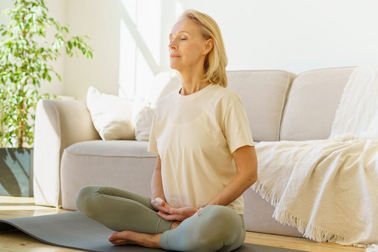Peaceful senior woman in lotus position meditation with closed eyes at home