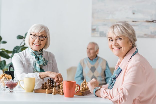 senior women playing chess and drinking coffee while senior man sitting on sofa senior women playing chess and drinking coffee while senior man sitting on sofa