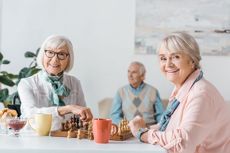 senior women playing chess and drinking coffee while senior man sitting on sofa