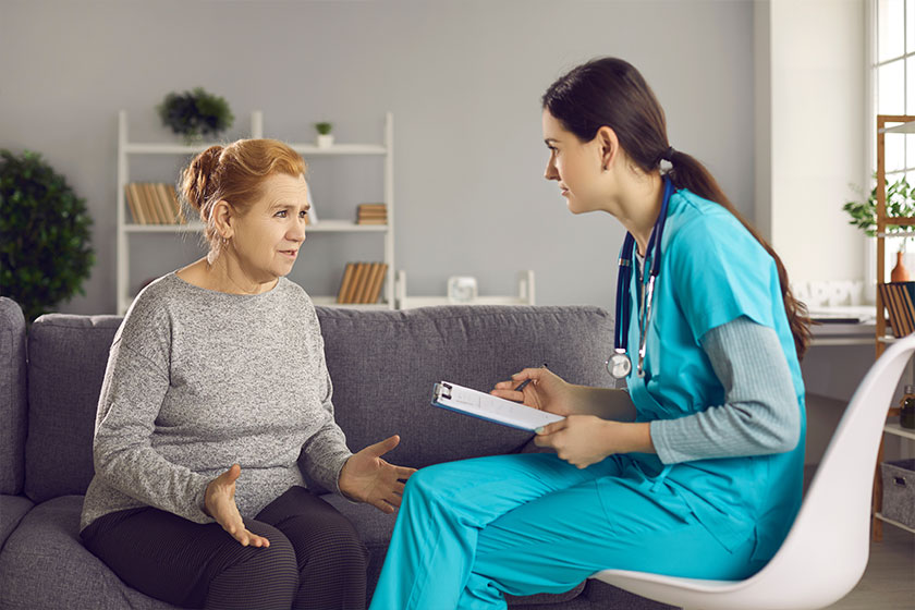 Young general practitioner with clipboard listening to senior female patient during home visit