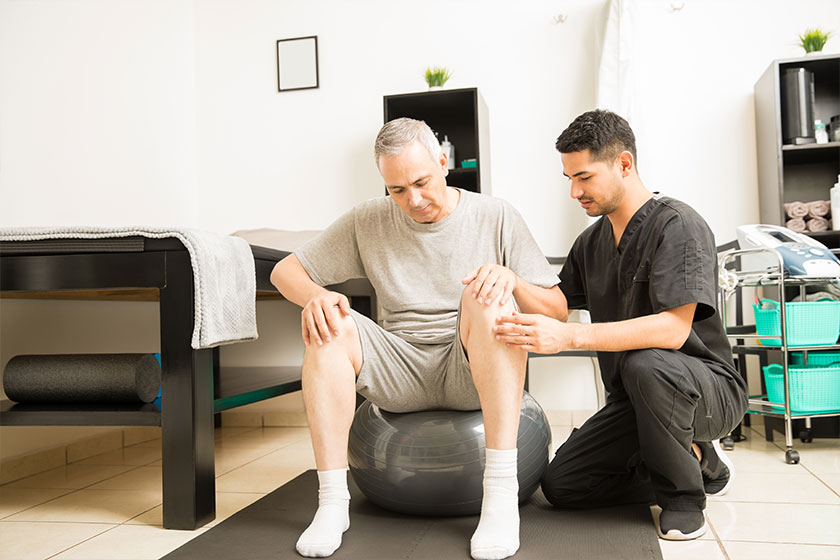 Therapist Assisting Mature Male Patient Sitting On Exercise Ball Therapist Assisting Mature Male Patient Sitting On Exercise Ball