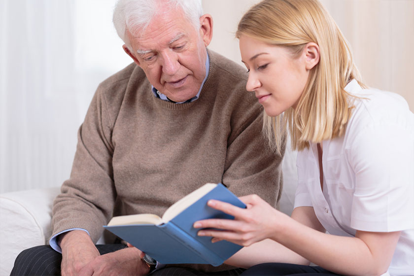 Young blonde volunteer reading book to senior man