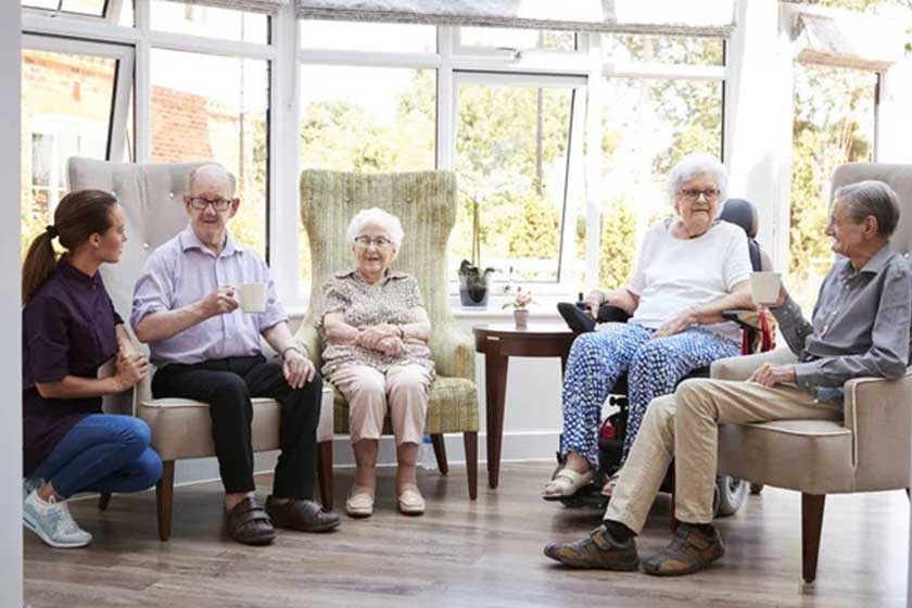 Male And Female Residents Sitting In Chairs And Talking With Carer In Lounge Of Retirement Home Male And Female Residents Sitting In Chairs And Talking With Carer In Lounge Of Retirement Home
