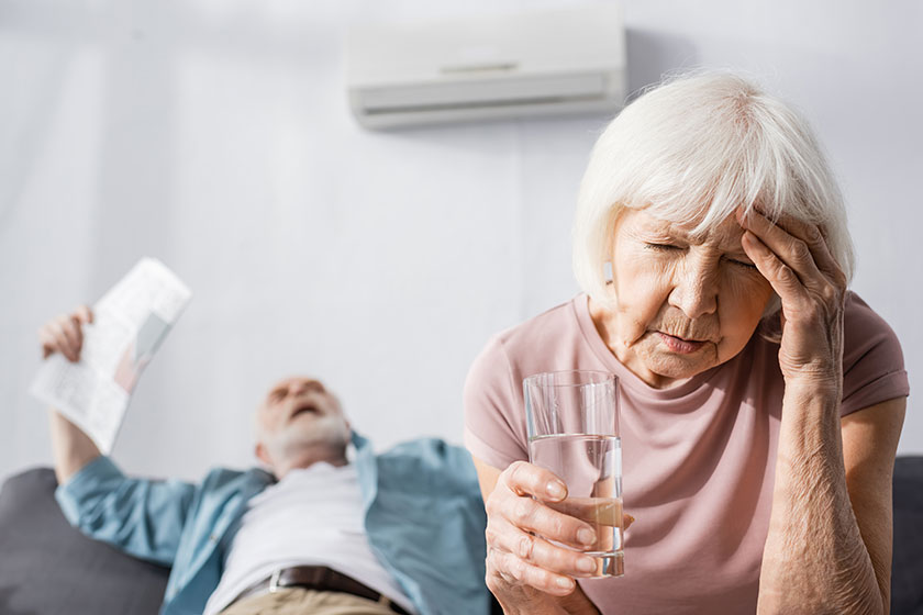 Selective focus of tired senior woman holding glass of water while feeling hot near husband with newspaper at home