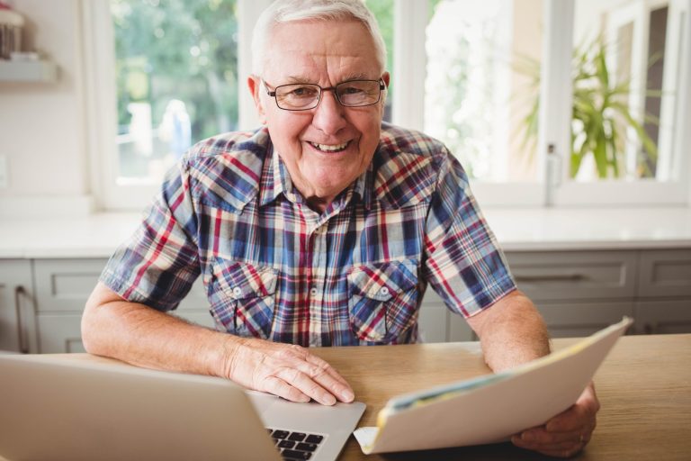 Happy senior man holding a document while using laptop at home
