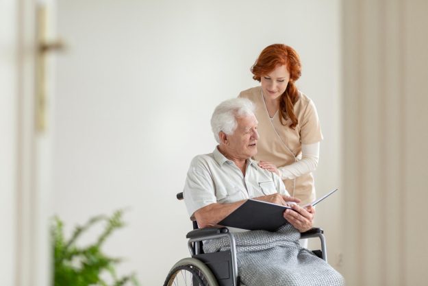 Caregiver,Supporting,Paralyzed,Elderly,Man,In,A,Wheelchair,With,Photo