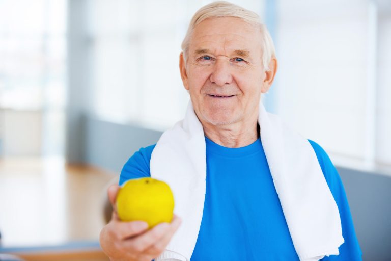 Join healthy lifestyle! Happy senior man with towel on shoulders stretching out green apple while standing in health club