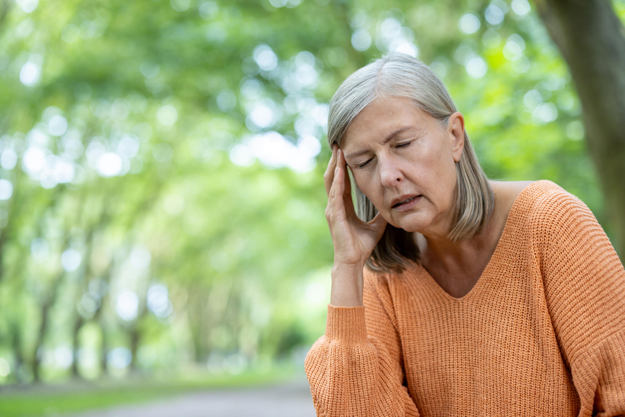 Senior woman experiencing stress with headache in green park, wearing orange sweater, showing discomfort