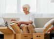 elderly woman sits on a sofa at home with boxes. collecting things with memories albums with photos and photo frames moving to a new place cleaning things and a happy smile. Lifestyle retirement.