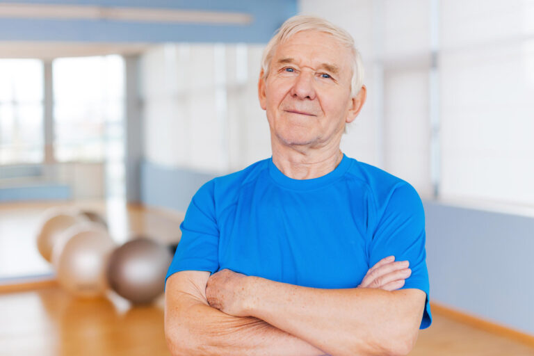 Confident and healthy. Confident senior man keeping arms crossed and looking at camera while standing in health club with sports equipment laying in the background