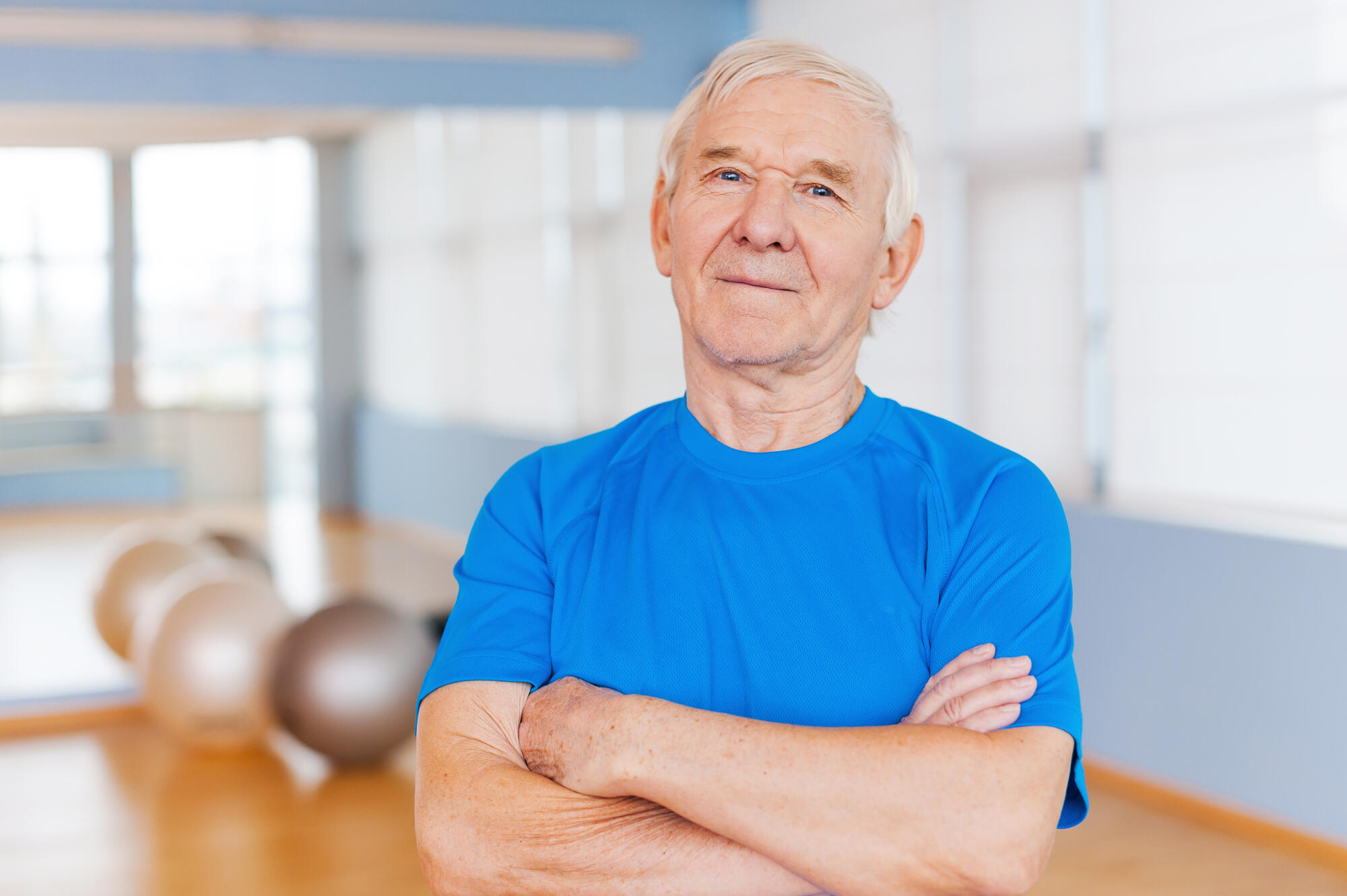 Confident and healthy. Confident senior man keeping arms crossed and looking at camera while standing in health club with sports equipment laying in the background Confident and healthy. Confident senior man keeping arms crossed and looking at camera while standing in health club with sports equipment laying in the background
