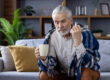 Sad senior gray haired man sitting on sofa at home and suffering from illness. Covered with a blanket, holding a cup of hot drink and a napkin