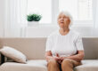 Elderly woman sits on sofa at home, bright spacious interior in old age smile, lifestyle. Grandmother with gray hair in a white T shirt and beige trousers.