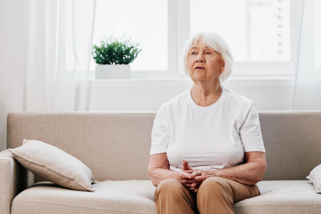 Elderly woman sits on sofa at home, bright spacious interior in old age smile, lifestyle. Grandmother with gray hair in a white T shirt and beige trousers.