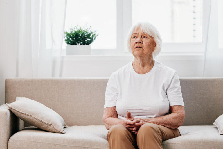 Elderly woman sits on sofa at home, bright spacious interior in old age smile, lifestyle. Grandmother with gray hair in a white T shirt and beige trousers.