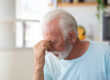 Tired, depressed senior man sitting on couch in living room feeling hurt and lonely. Aged, white haired man touching forehead suffering from severe headache or recalling bad memories