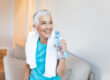 Elderly woman sitting on the sofa, exhausted after the daily training.Senior woman taking a break while exercising at home. Athletic mature woman holding bottle of water,Having a towel around her neck